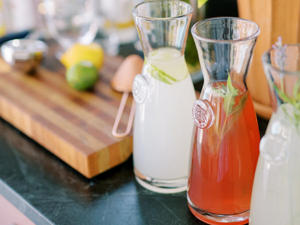 Pomegranate Lemonade in a pitcher with a large bottle of Sonoma Syrup Co. Pomegranate Simple Syrup and 2 glasses with lemon slices