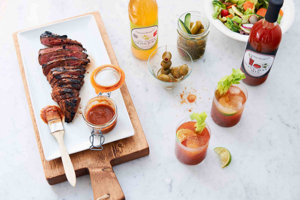Photo of Barbecue meat on a cutting board with Bloody Olive Mary Tri Tip BBQ Sauce. Paired with two Bloody Mary cocktails and a fresh salad.