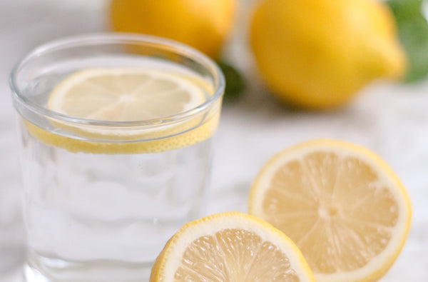 Photo of a Lemon Extract bottle on a bench next to a glass with sparkling water being poured into it. A vibrant lemon tree sits in the background.