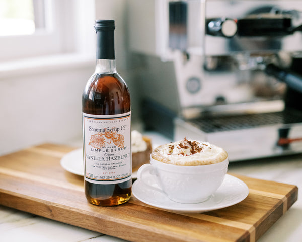 Photo of Vanilla Hazelnut Simple Syrup next to a Hazelnut Infused Latte recipe topped with cocoa powder next to an espresso machine.