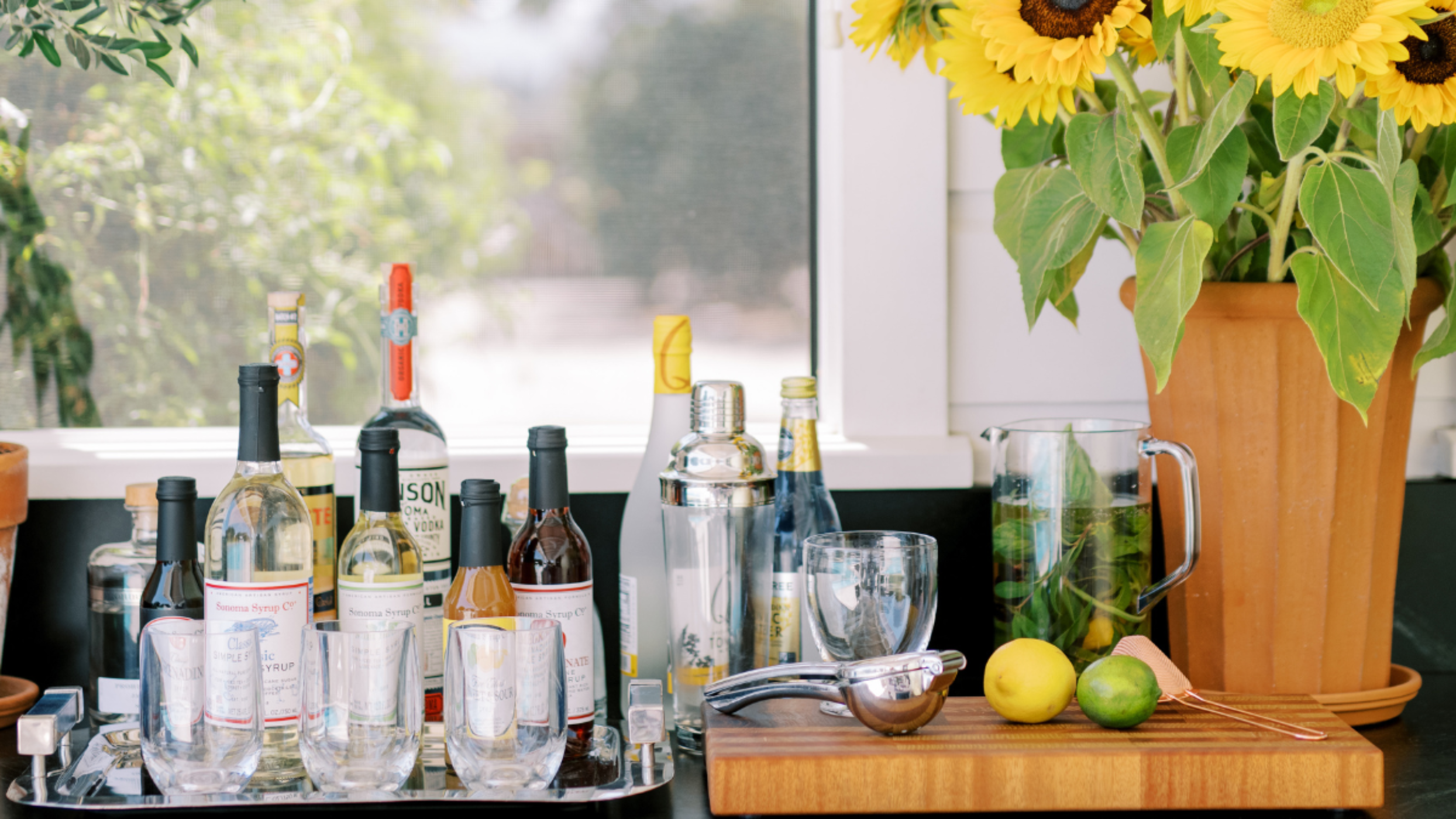 Beverage setup with bottles, glasses, and a vase of sunflowers on a counter.