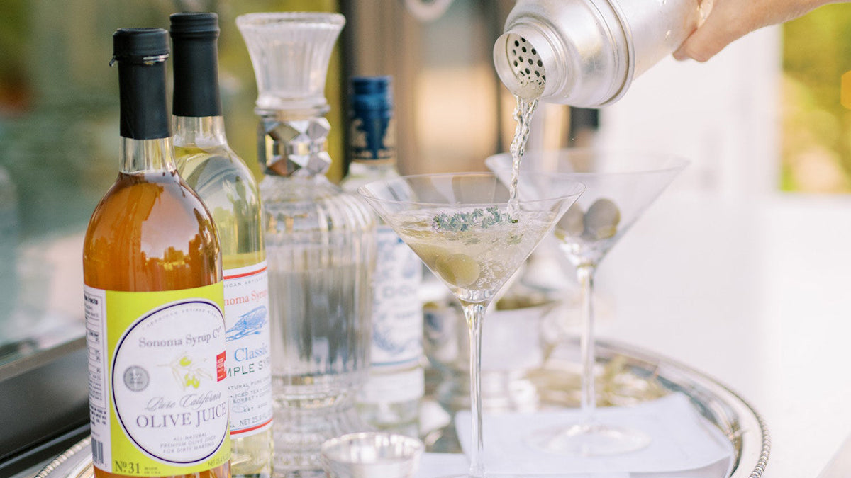 Mixologist pouring dirty martini into glass on tray with bottles of Sonoma Syrup Co olive juice and simple syrup
